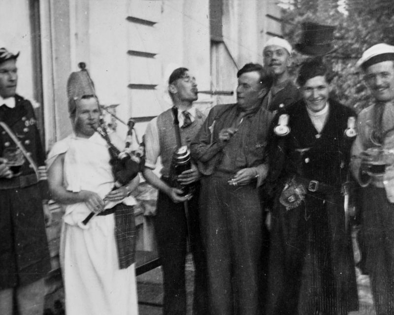 A number of men in fancy dress standing outside a large white building, one of the men is playing some bagpipes and some of the others are holding glasses of beer.