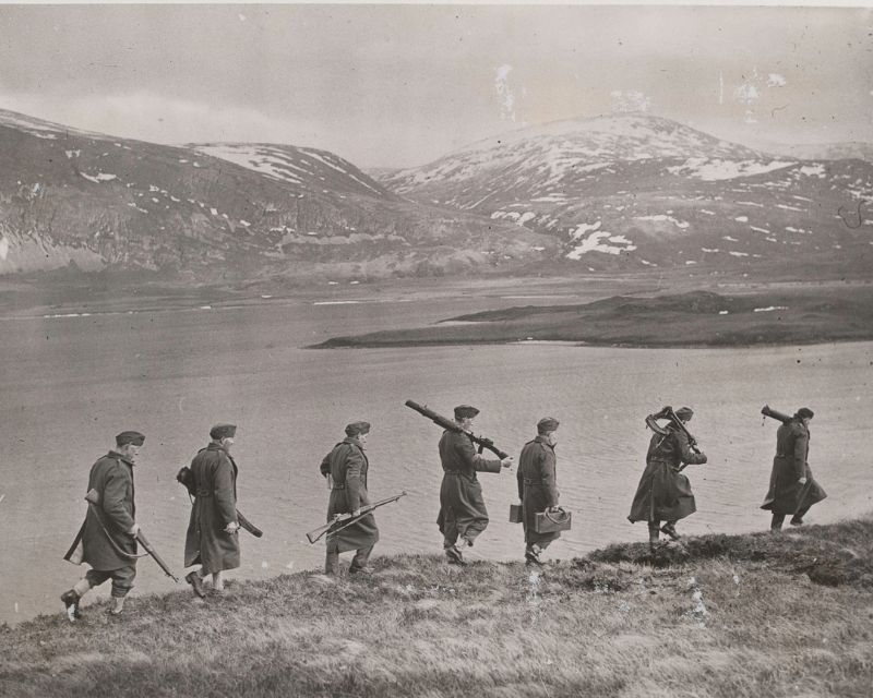 A line of Home Guard soldiers carrying guns and equipment along a hillside, mountains in the background.