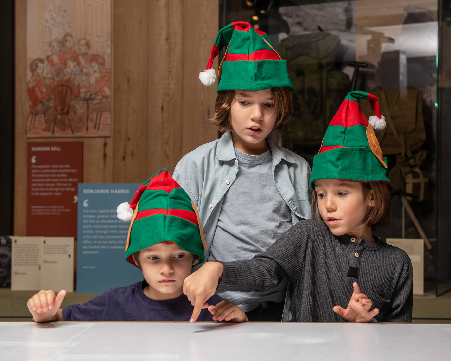 Three children wearing Christmas hats, exploring the collection via a touch screen